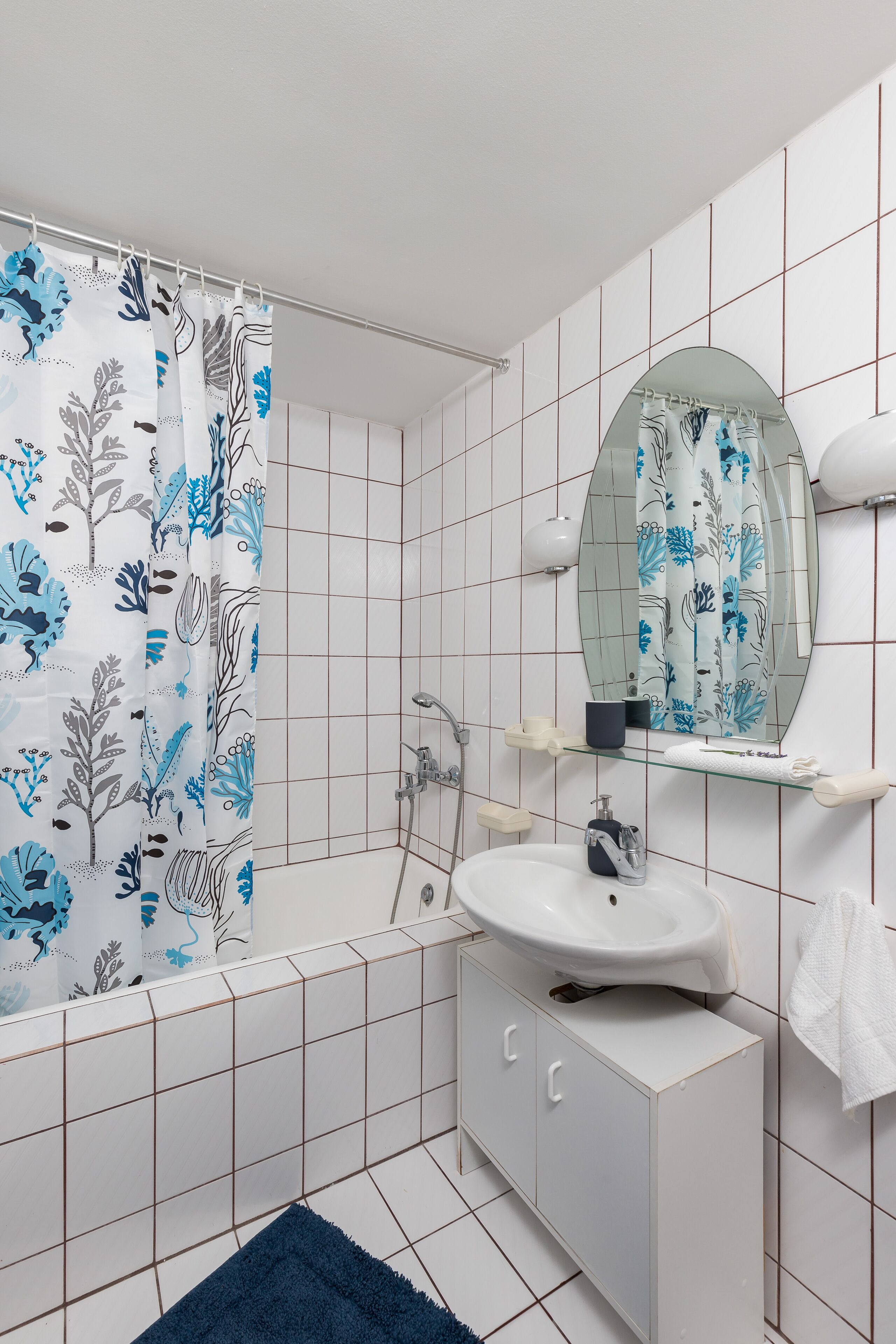 Une salle de bain avec des murs recouverts de carreaux blancs, un lavabo blanc, un miroir et un rideau de douche à motifs floraux.
