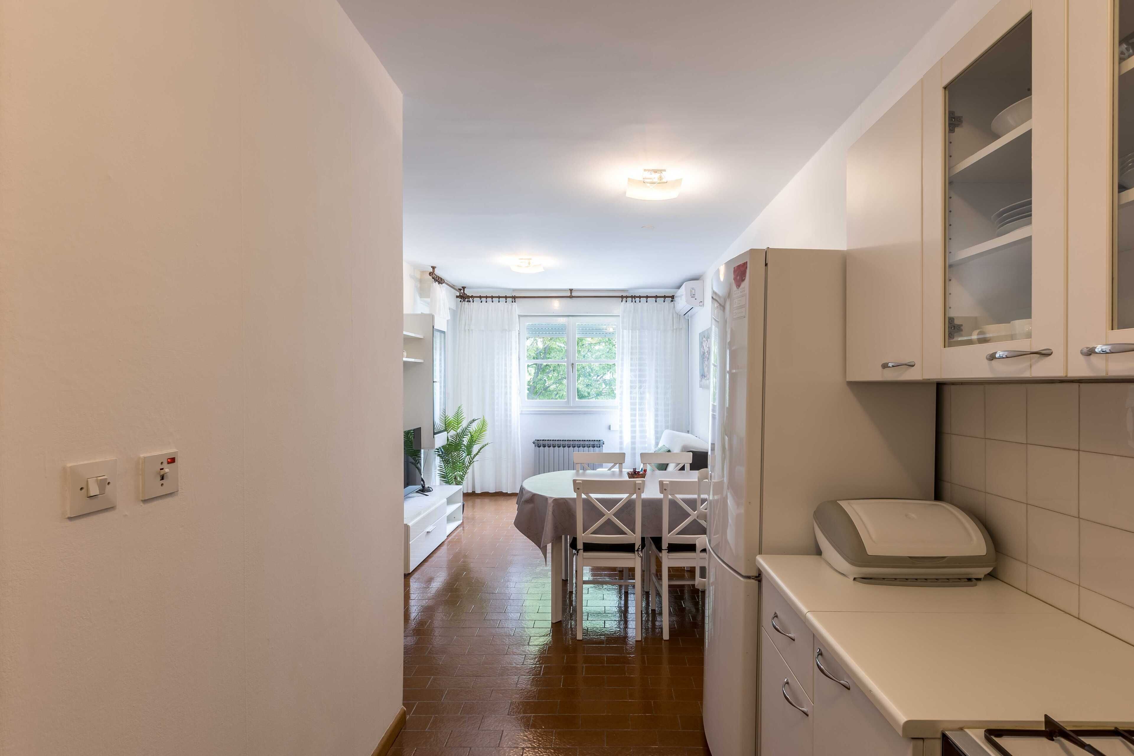 A kitchen with white cabinets, a dining table, and chairs. There is a window with curtains, a radiator, and a plant.