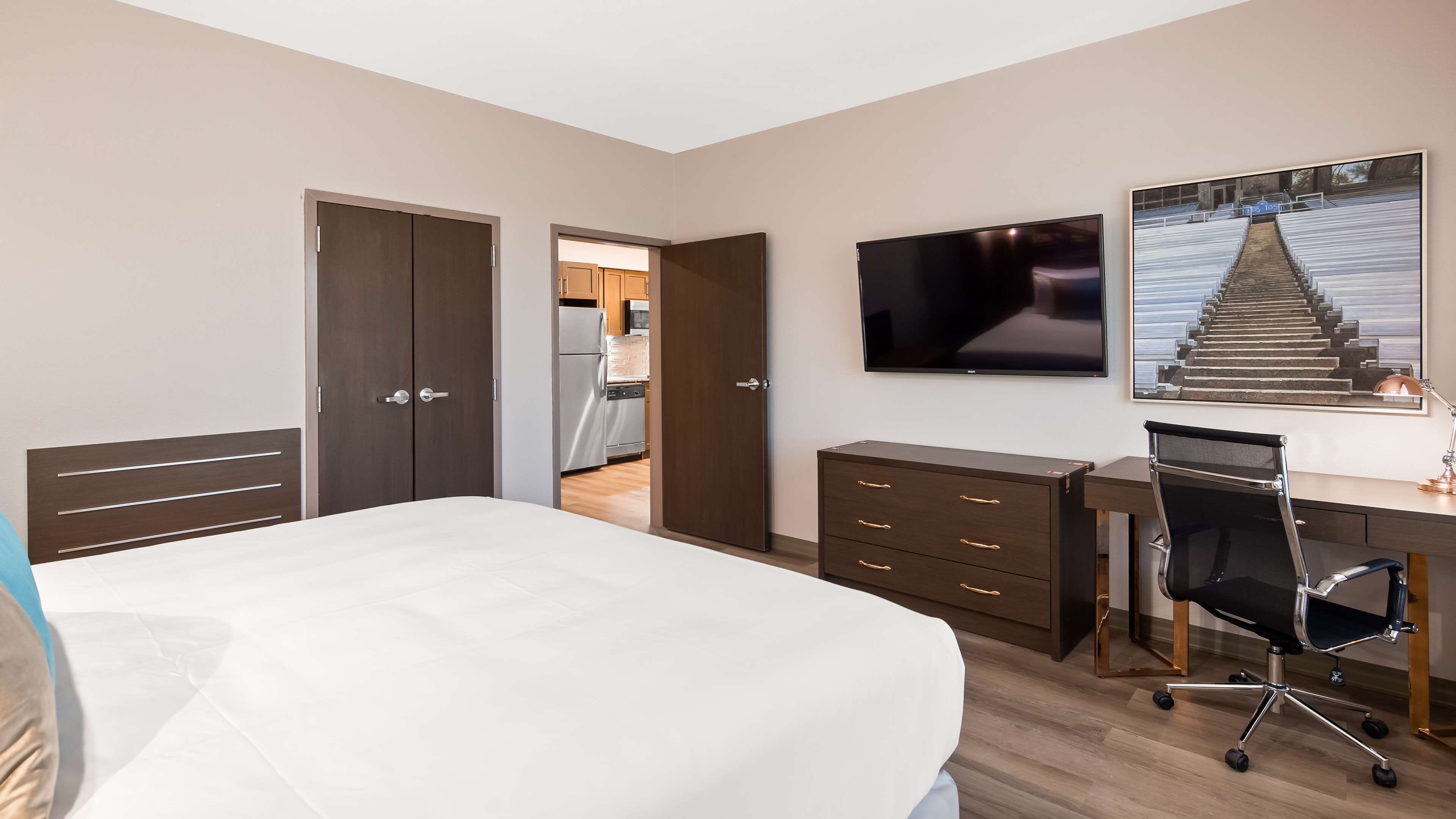 A modern hotel room with a large bed, a desk with a chair, a flat-screen TV, and a staircase leading to the kitchen.