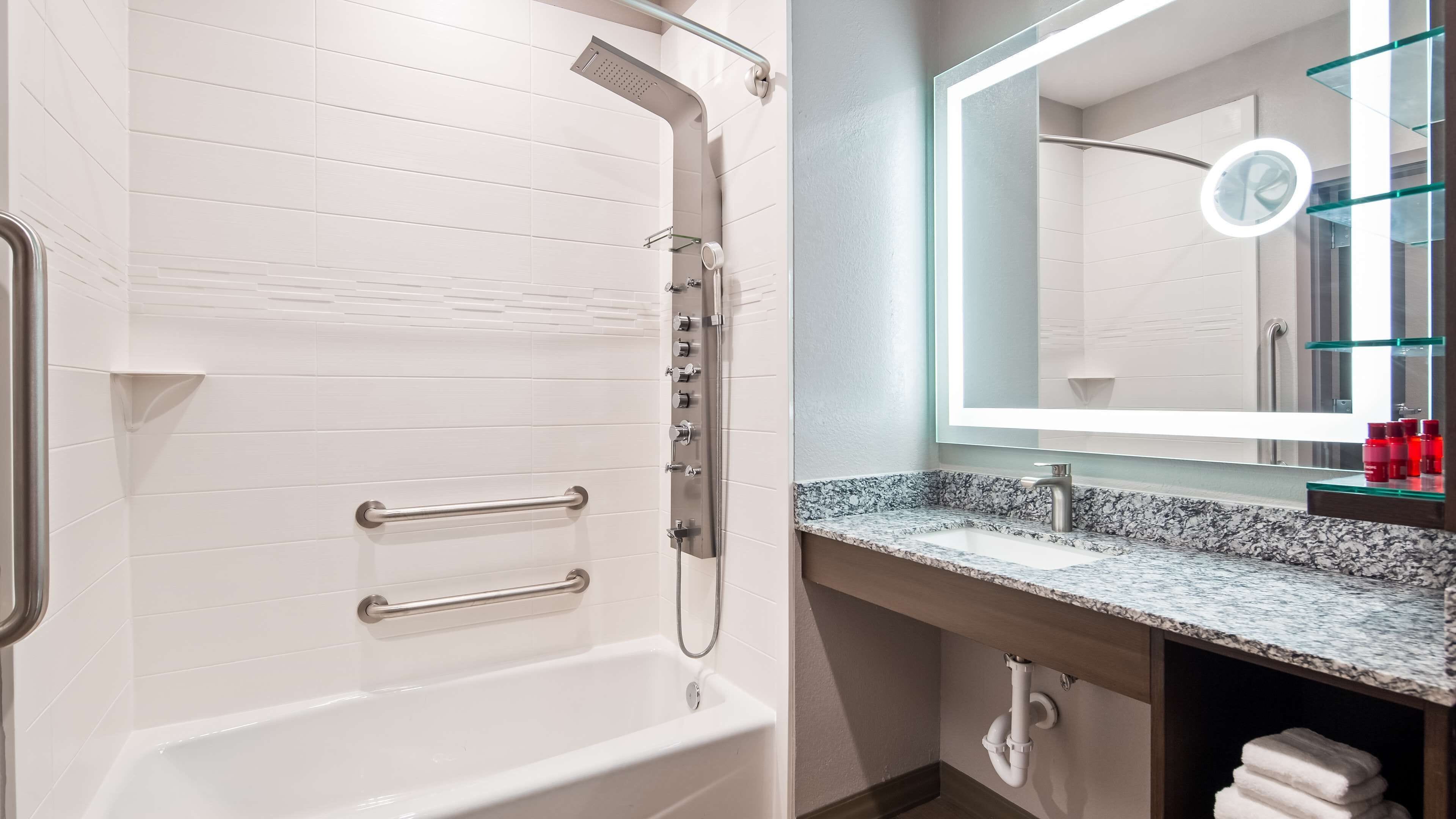 A modern bathroom with a white tiled shower, a glass-framed mirror, a marble countertop, and a visible towel stack.