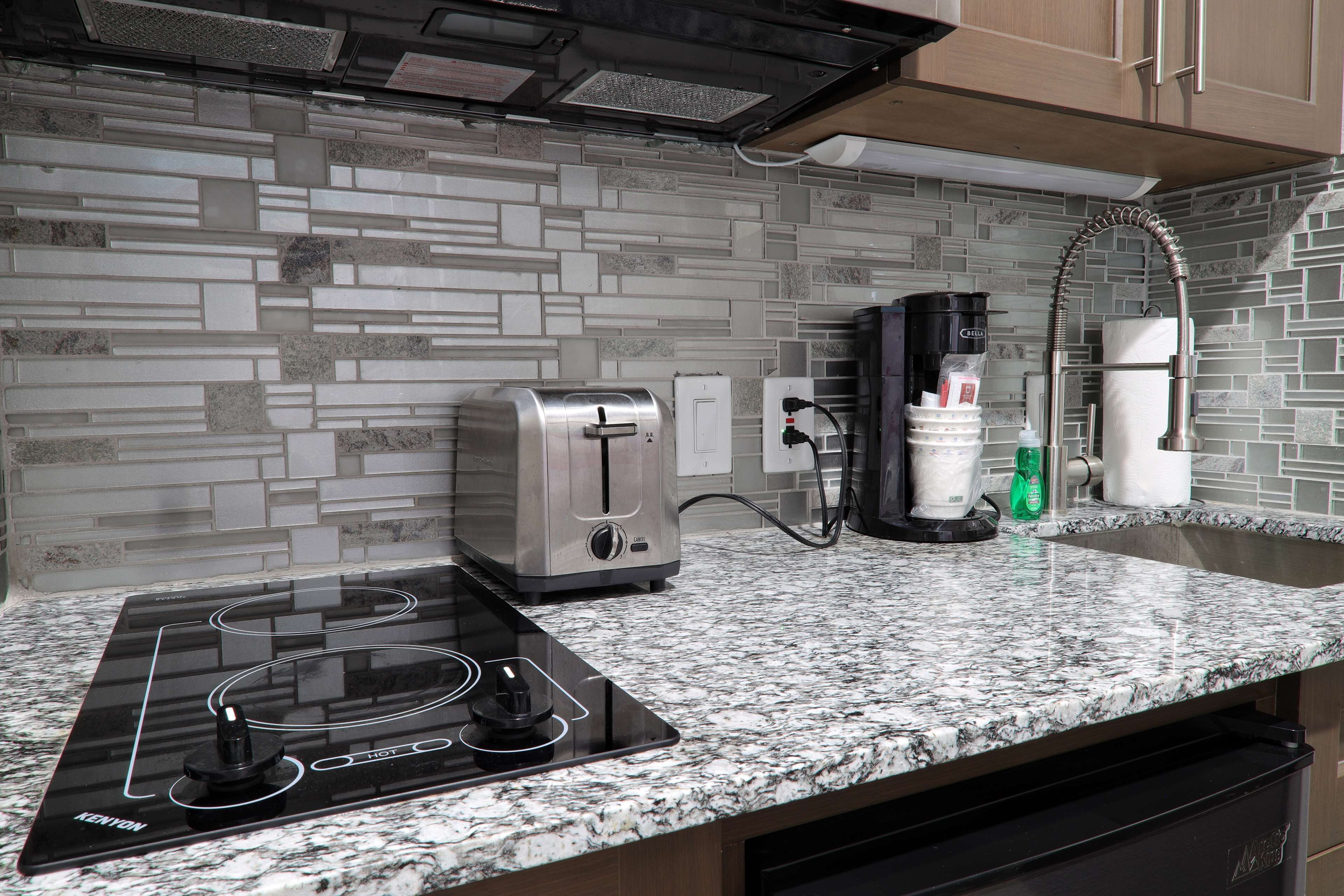 A kitchen with a granite countertop, a Kenyon induction cooktop, a toaster, a coffee maker, and a dishwasher.