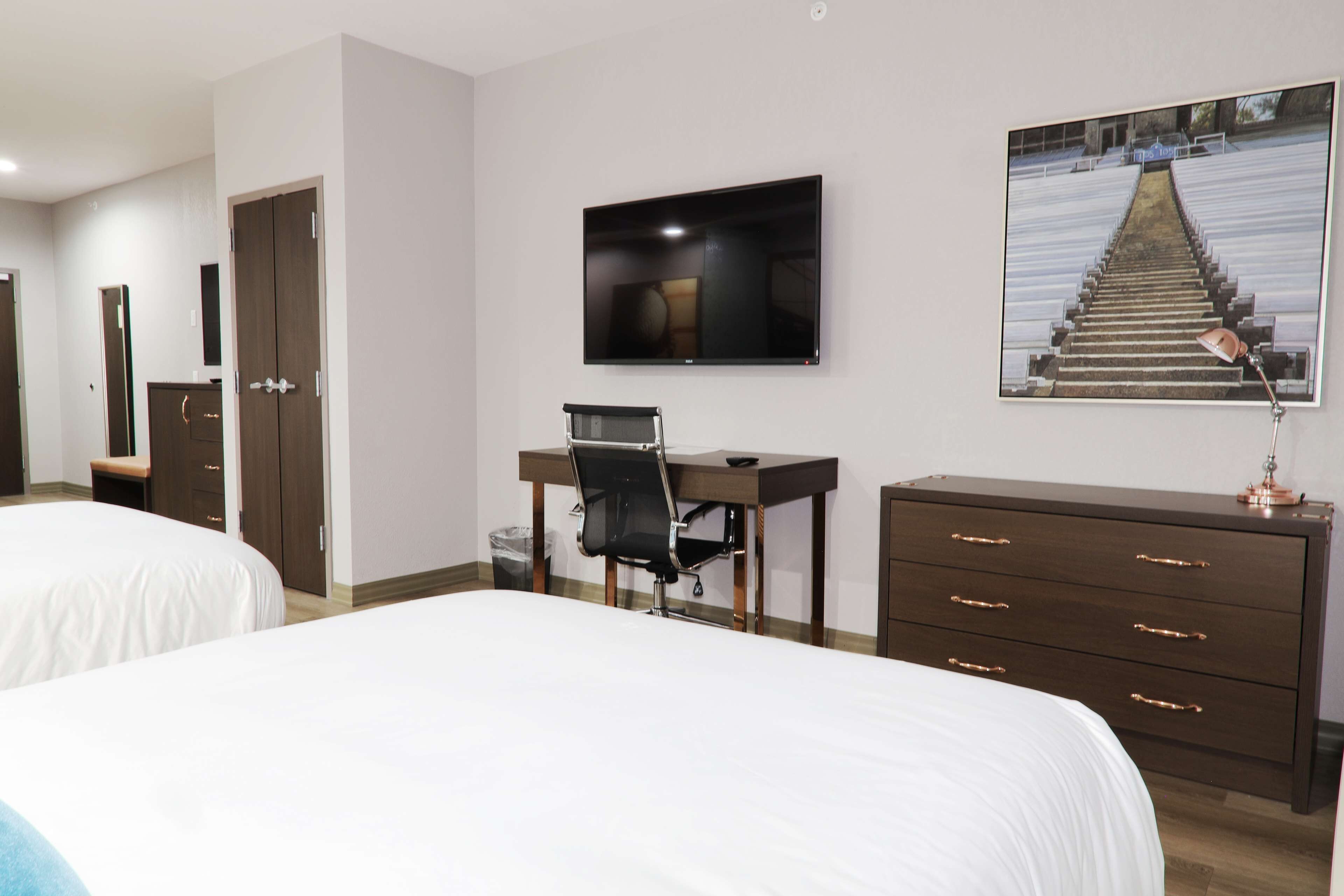 Ein Hotelzimmer mit zwei Betten, einem Schreibtisch, einem Sessel, einem Fernseher, einem Schminktisch und einem gerahmten Bild an der Wand.