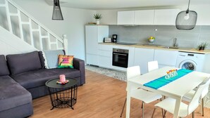Un apartamento moderno con una sala de estar que incluye un sofá gris oscuro, una pequeña mesa redonda de centro y una mesa de comedor blanca con sillas. Se ve una escalera con barandilla blanca al fondo.