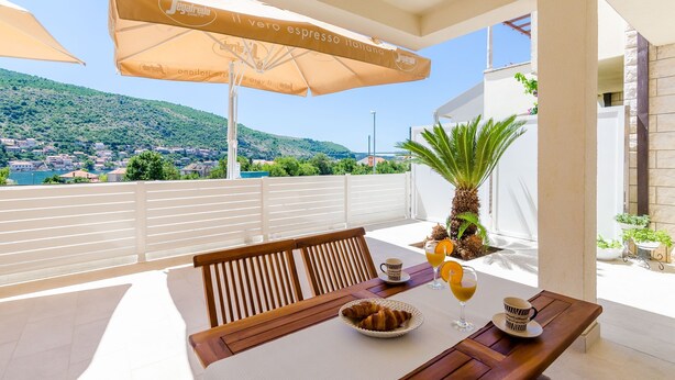 A terrace with a table set for two, a wooden bench, and a view of a coastal town. - Apartment Moon River (Dubrovnik)