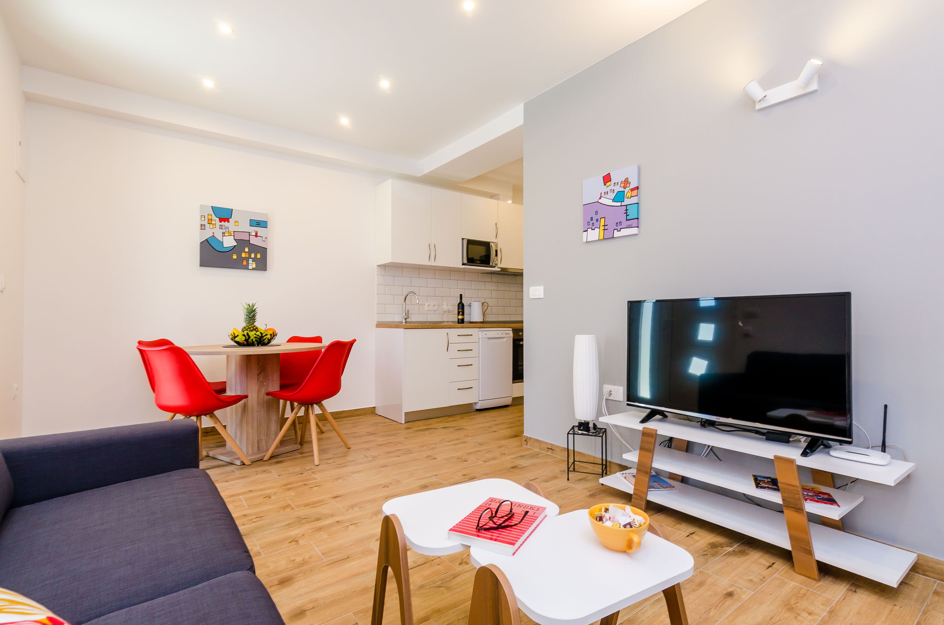 Un salon moderne avec un téléviseur à écran plat, un canapé, une table à manger avec des chaises rouges, et un coin cuisine à l’arrière-plan.