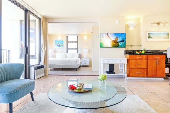 A modern hotel room with a glass coffee table, a sofa, a bed, a kitchen area, and a large window.