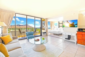 A modern hotel room with a balcony, a sofa, a coffee table, a TV, and a kitchen area.