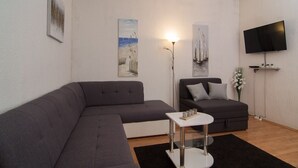 Un salon moderne avec un canapé d’angle gris, un canapé gris plus petit, une table basse blanche et un téléviseur à écran plat fixé au mur.