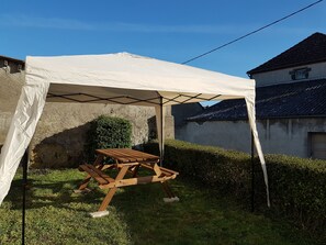 A wooden picnic table under a white canopy in a grassy outdoor area with a stone wall and a building in the background.