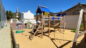 A playground with a slide and climbing structures, surrounded by sand, with buildings in the background. - Gite in the heart of an artisan village - marsh views - near ocean (Sallertaine)