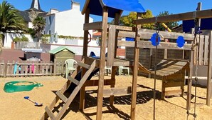 Una struttura giochi in legno con tetto blu e uno scivolo, su terreno sabbioso con un edificio bianco e palme sullo sfondo.