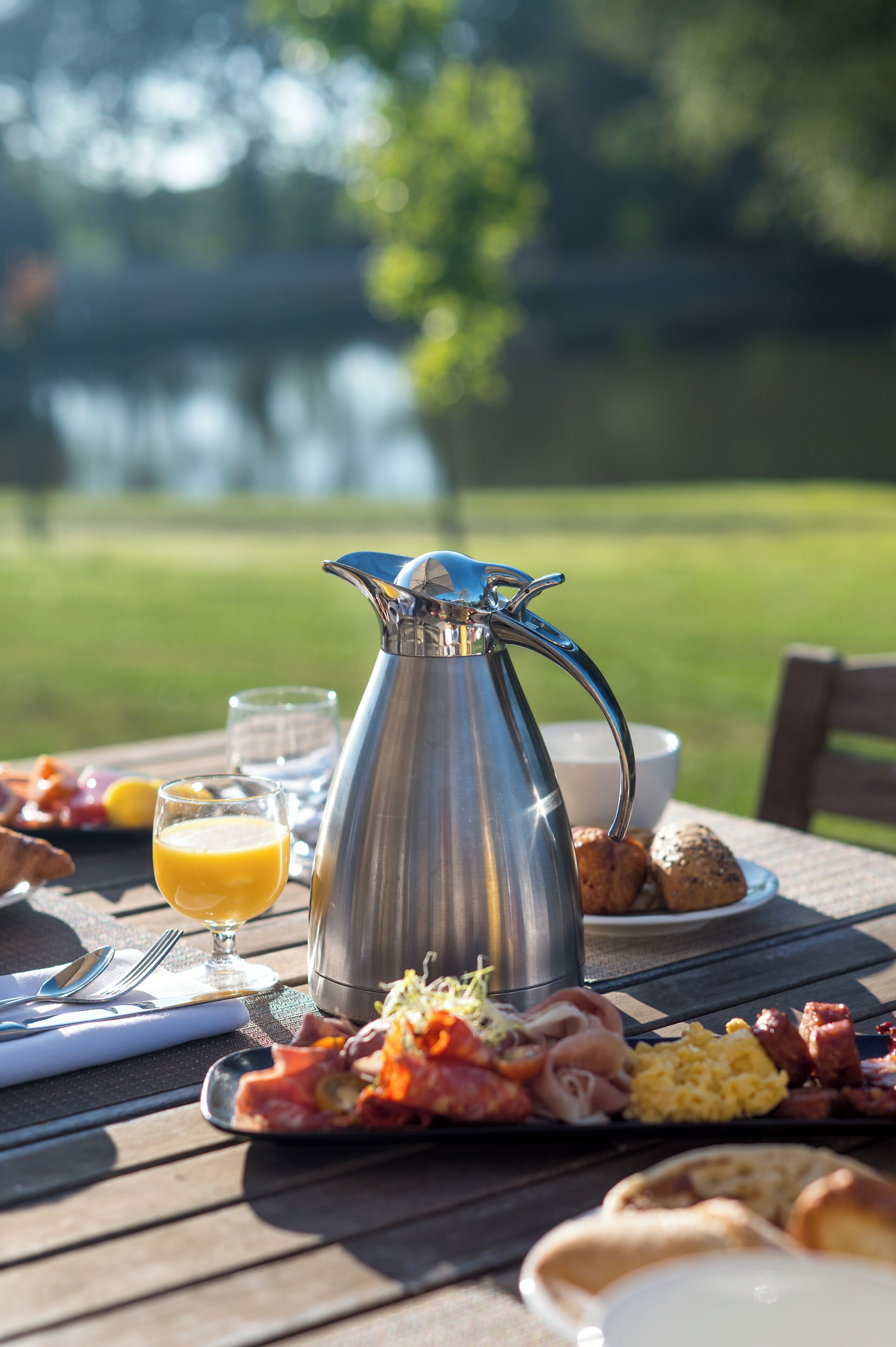 A breakfast setting with a metal pitcher, glasses of orange juice, croissants, and a variety of meats and eggs on a wooden table.