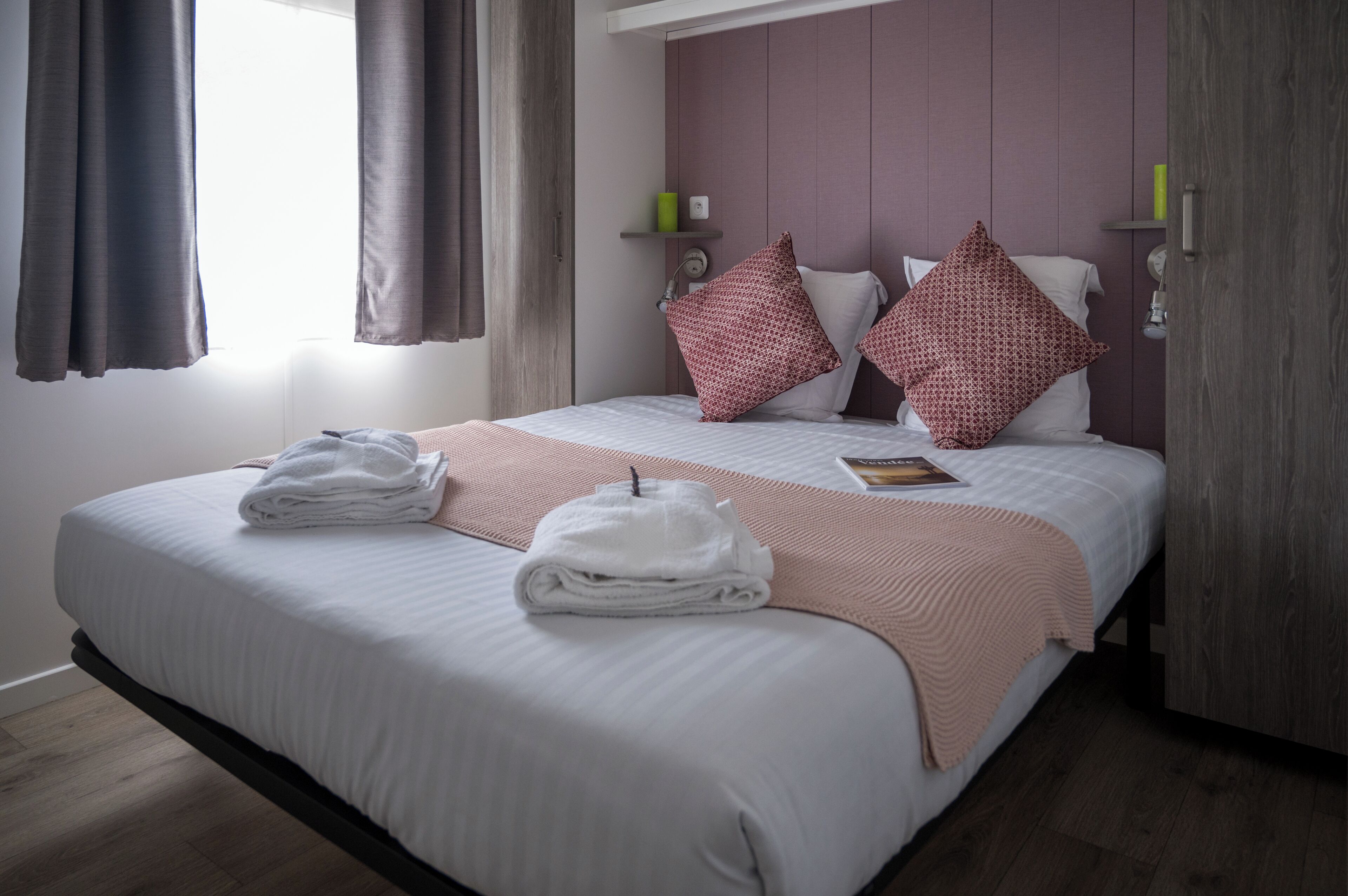 A neatly made bed with towels, a pink throw blanket, and two red patterned pillows.