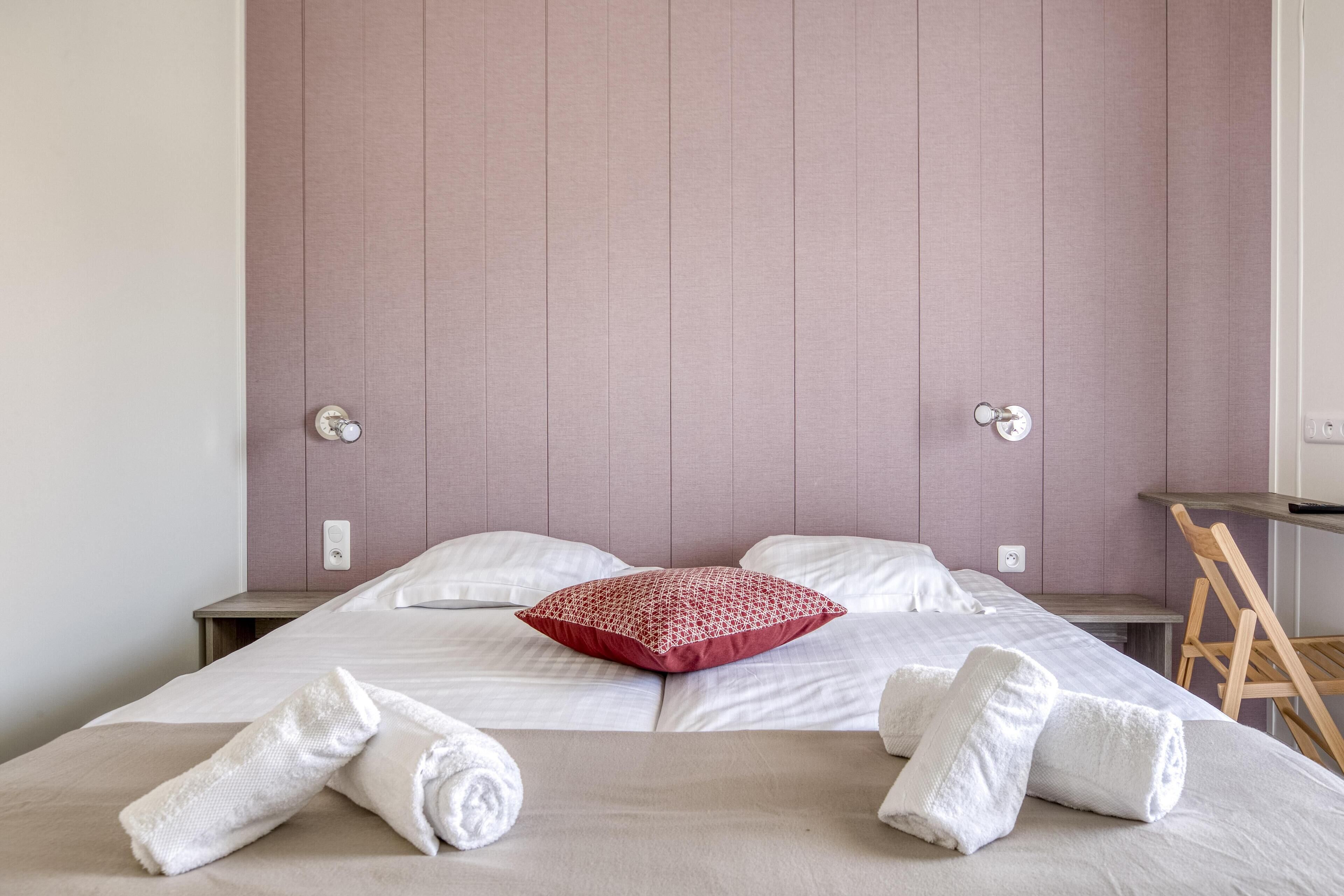 Una cama bien hecha con sábanas blancas, una almohada roja estampada y dos toallas blancas enrolladas.
