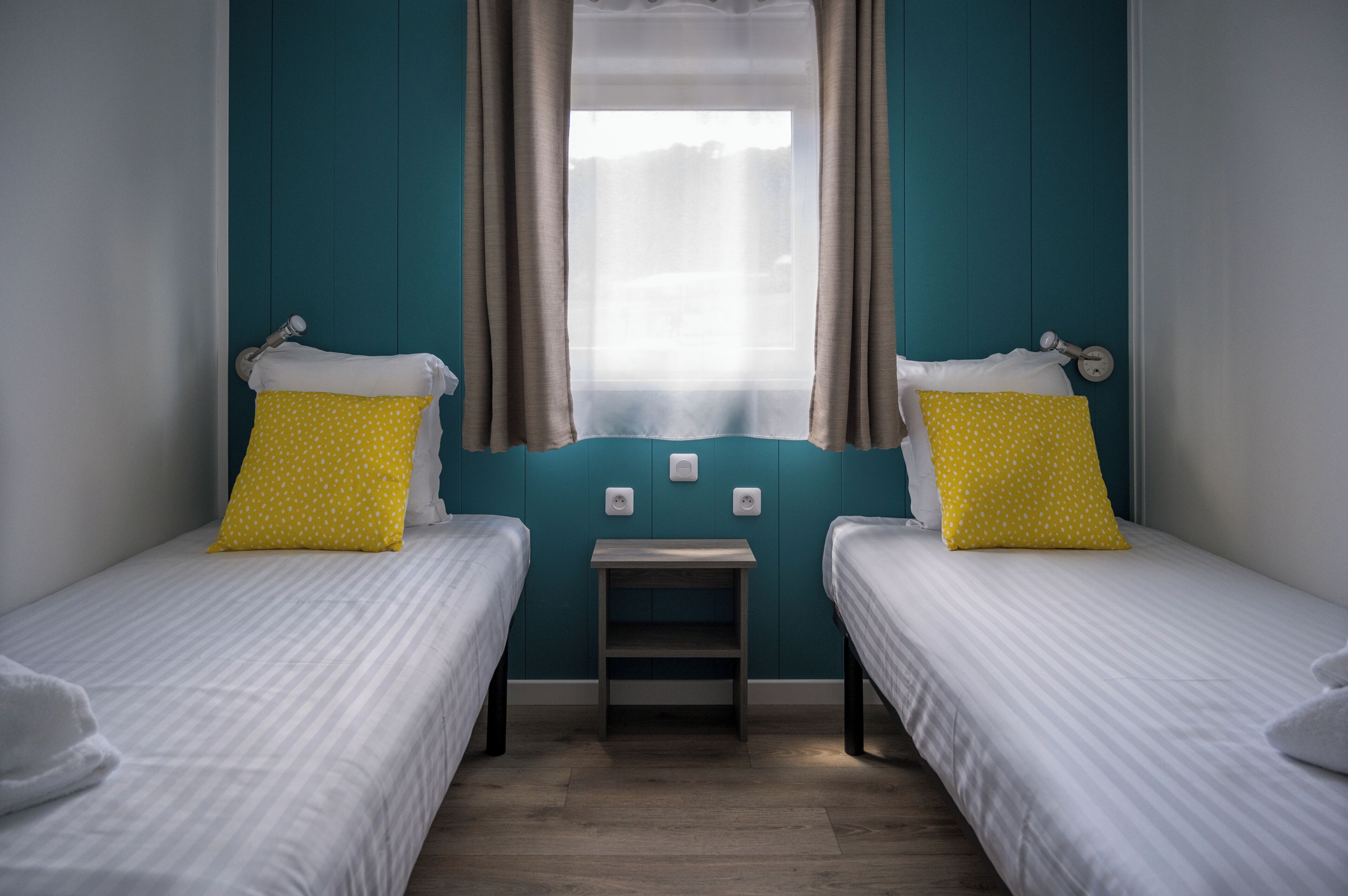 A twin room with two single beds, each with a white bedspread and a yellow pillow with white polka dots. There is a small wooden nightstand between the beds.