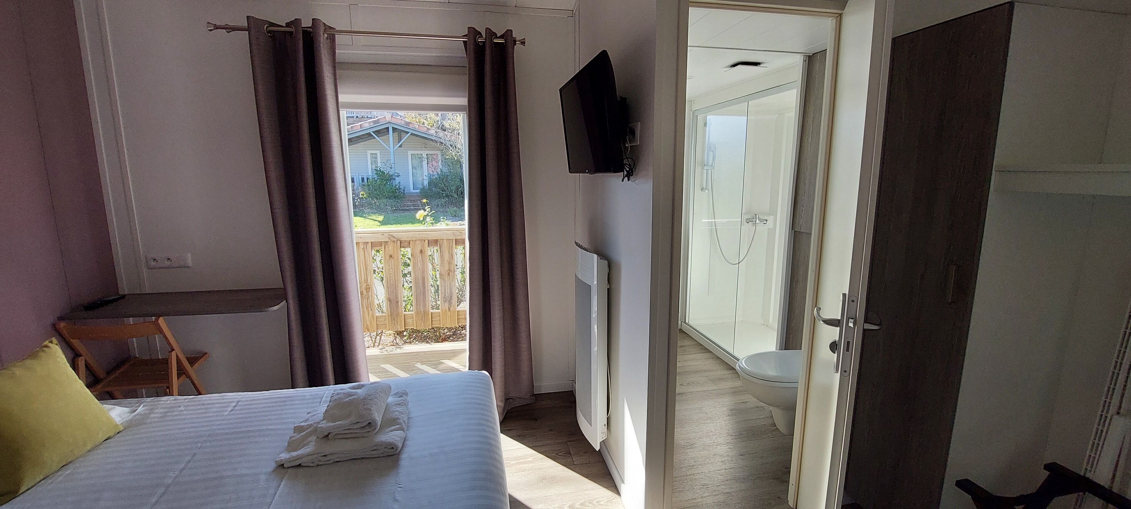 Un dormitorio con cama, ventana con cortinas, una silla de madera, televisor y vistas a una casa y jardín.