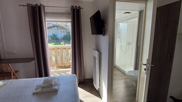 Un dormitorio con cama, ventana con cortinas, una silla de madera, televisor y vistas a una casa y jardín.