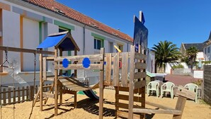 A wooden playground with slides and climbing structures, surrounded by a sandy area and a building with a tiled roof. - Gîte in the heart of an artists' village - close to the ocean - marsh views (Sallertaine)