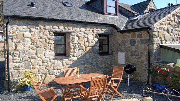 A stone building with a stone exterior, a wooden table and chairs, a barbecue grill, and a small garden area with potted plants.