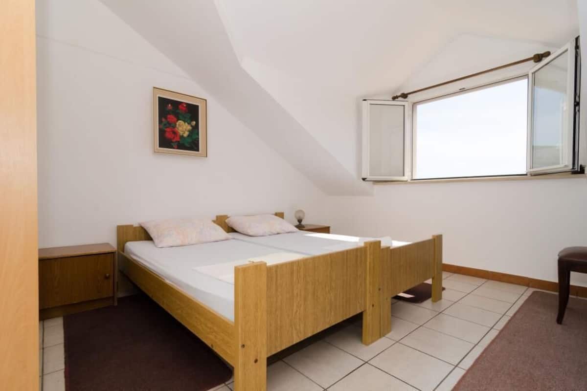 Une chambre individuelle avec un lit en bois, une table de chevet, une petite armoire, une fenêtre donnant sur un paysage agréable et un tableau au mur.
