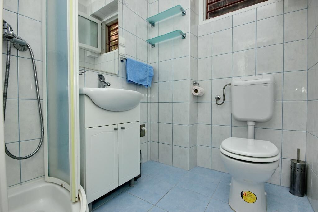 Et badeværelse med hvid vask, toilet og brusebad. Der er glas hylder over vasken, og et blåt håndklæde hænger på væggen.