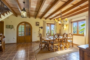 A dining area with a wooden table and chairs, a chandelier, and a view of the outdoors through large windows.