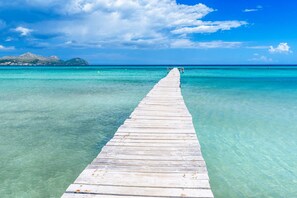 Un muelle de madera se adentra en aguas turquesas cristalinas, dirigiéndose hacia una tierra distante con montañas.