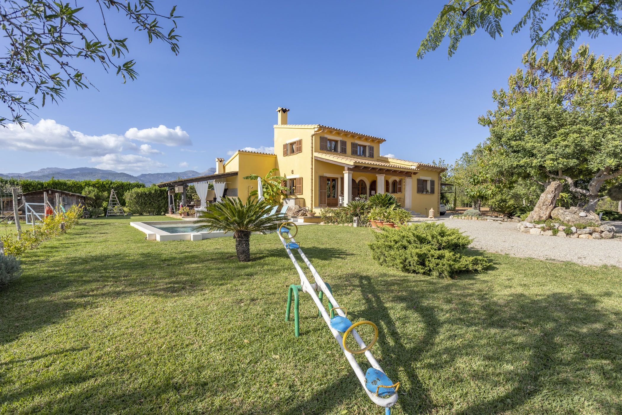 A yellow villa with a swimming pool, a seesaw, and a well-maintained garden.