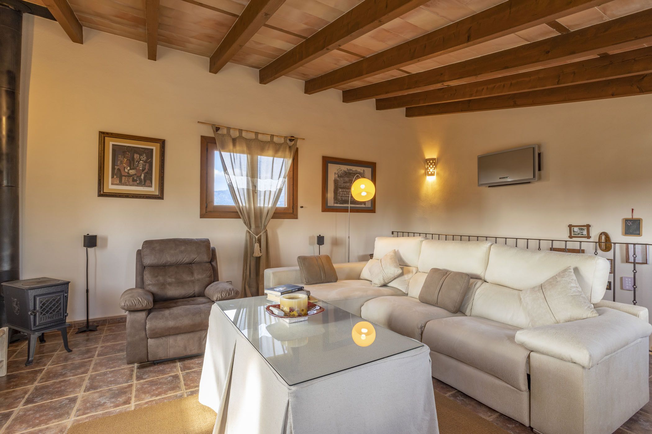 Una sala de estar con sofá, sillón y una mesa de centro de vidrio. Incluye una chimenea, un televisor montado en la pared y una ventana con cortinas.