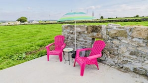 A patio with two bright pink chairs and a small round table, set against a stone wall with a view of a green field and distant buildings. - The Cuddly Cow Cosy Barn Studio Campview Farm Stay (Ballyshannon)