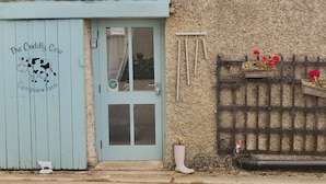 A rustic building with a blue door labeled "The Cuddly Cow Campview Farm," a wooden fence with flower baskets, and a small cow decoration on the wall. - The Cuddly Cow Cosy Barn Studio Campview Farm Stay (Ballyshannon)