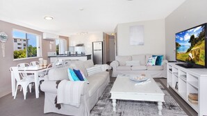 Una sala de estar moderna con un sofá blanco, una mesa de centro y un televisor. Se observa una cocina al fondo con armarios blancos y un refrigerador.