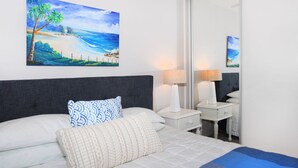 Habitación de hotel con cama, dos lámparas de noche, una mesita, un cuadro de una playa y un espejo grande.