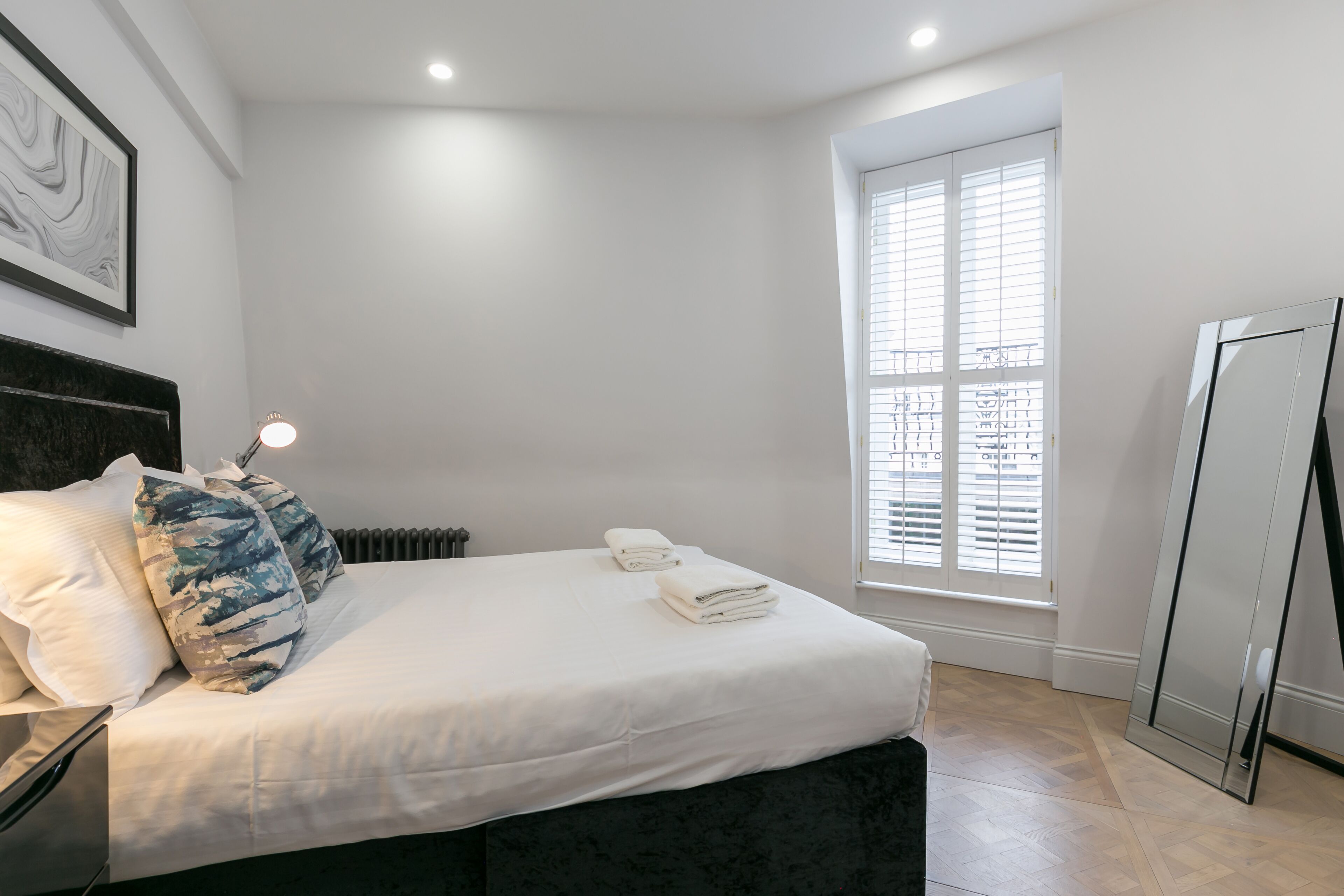 A bedroom with a bed, white bedding, a headboard, a window with blinds, and a radiator.