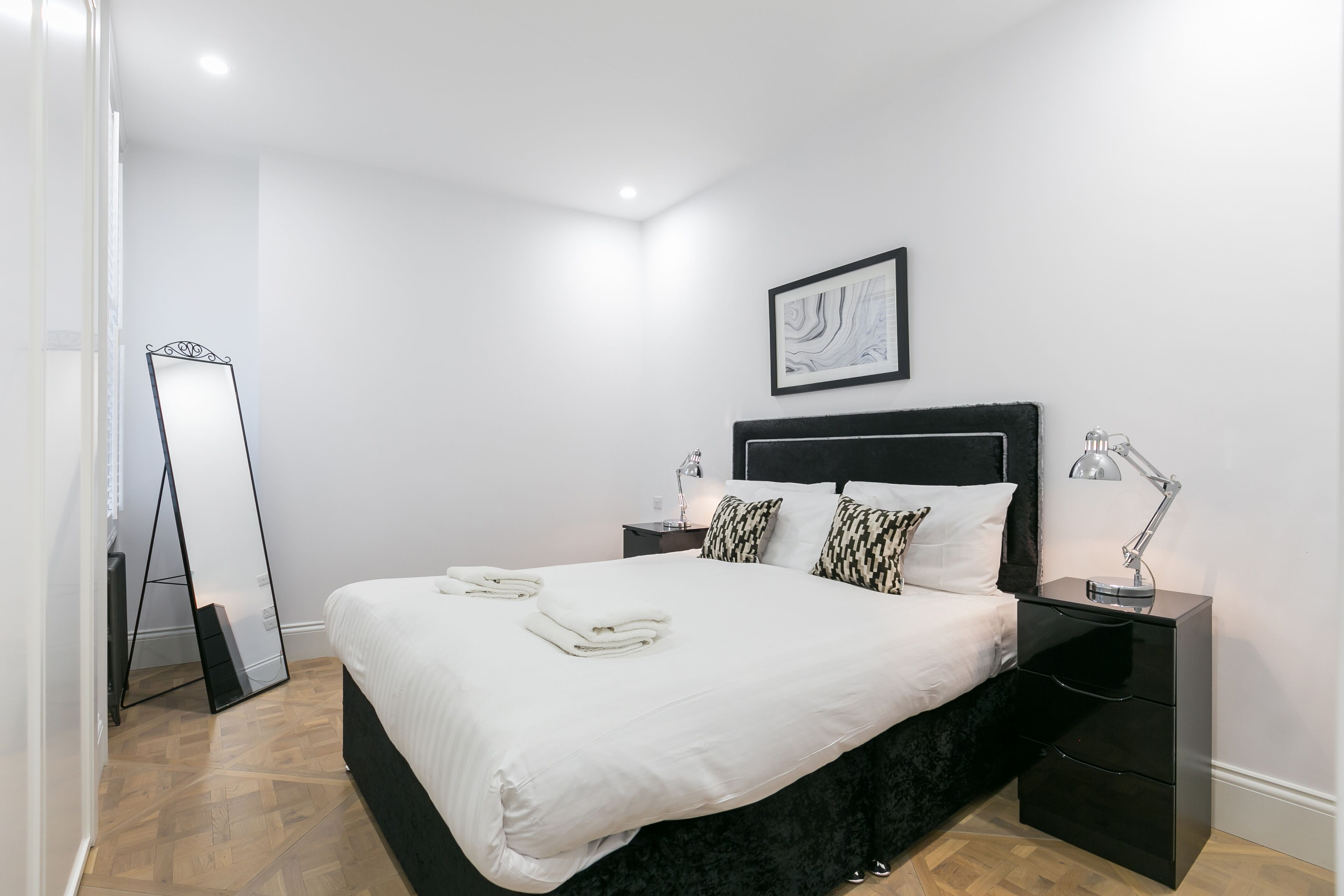 Ein ordentlich eingerichtetes Schlafzimmer mit einem großen Bett, einem Nachttisch mit Lampe, einem schwarzen Beistelltisch und einem großen Spiegel.