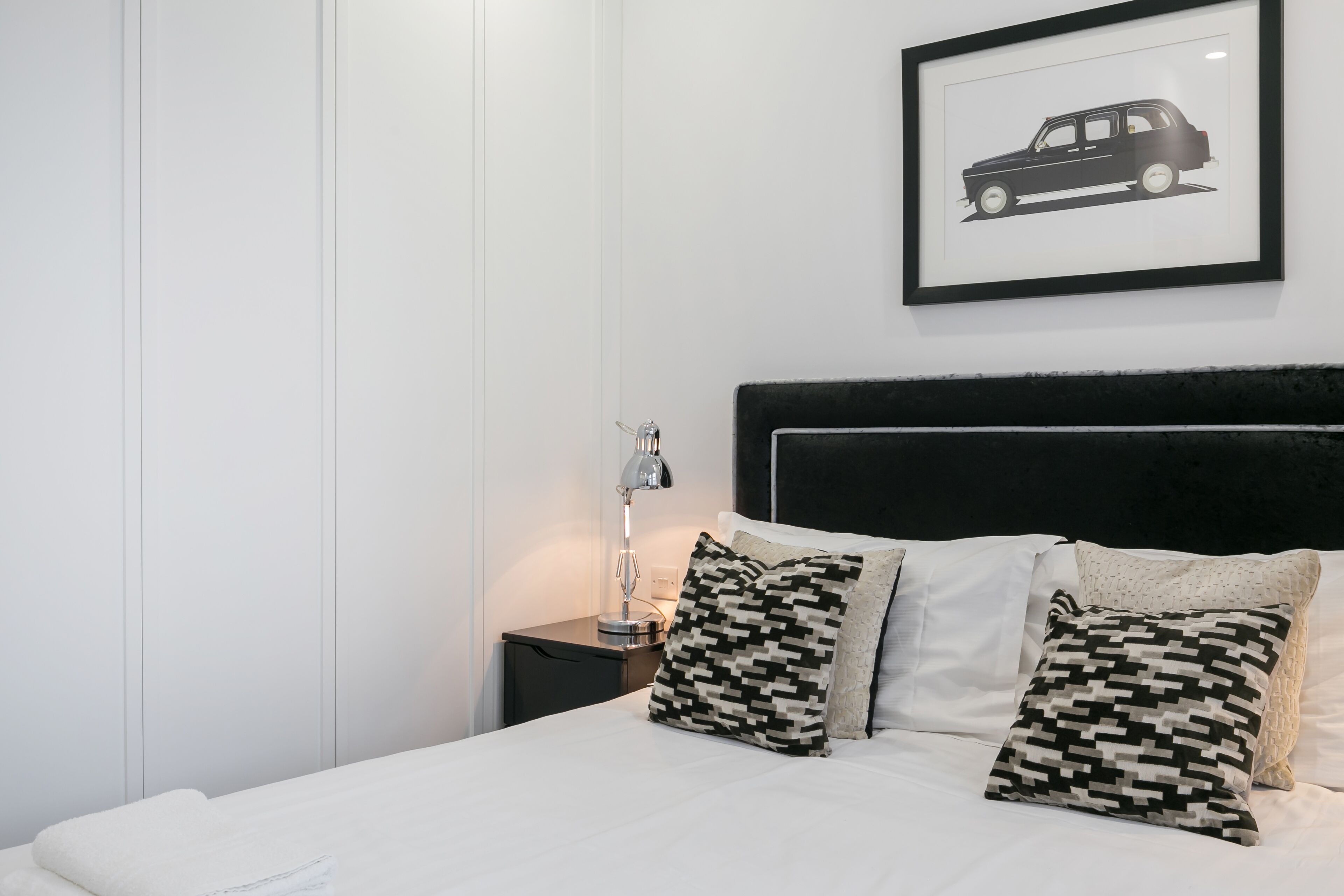 A neatly made bed with patterned pillows, a framed picture on the wall, and a bedside lamp.