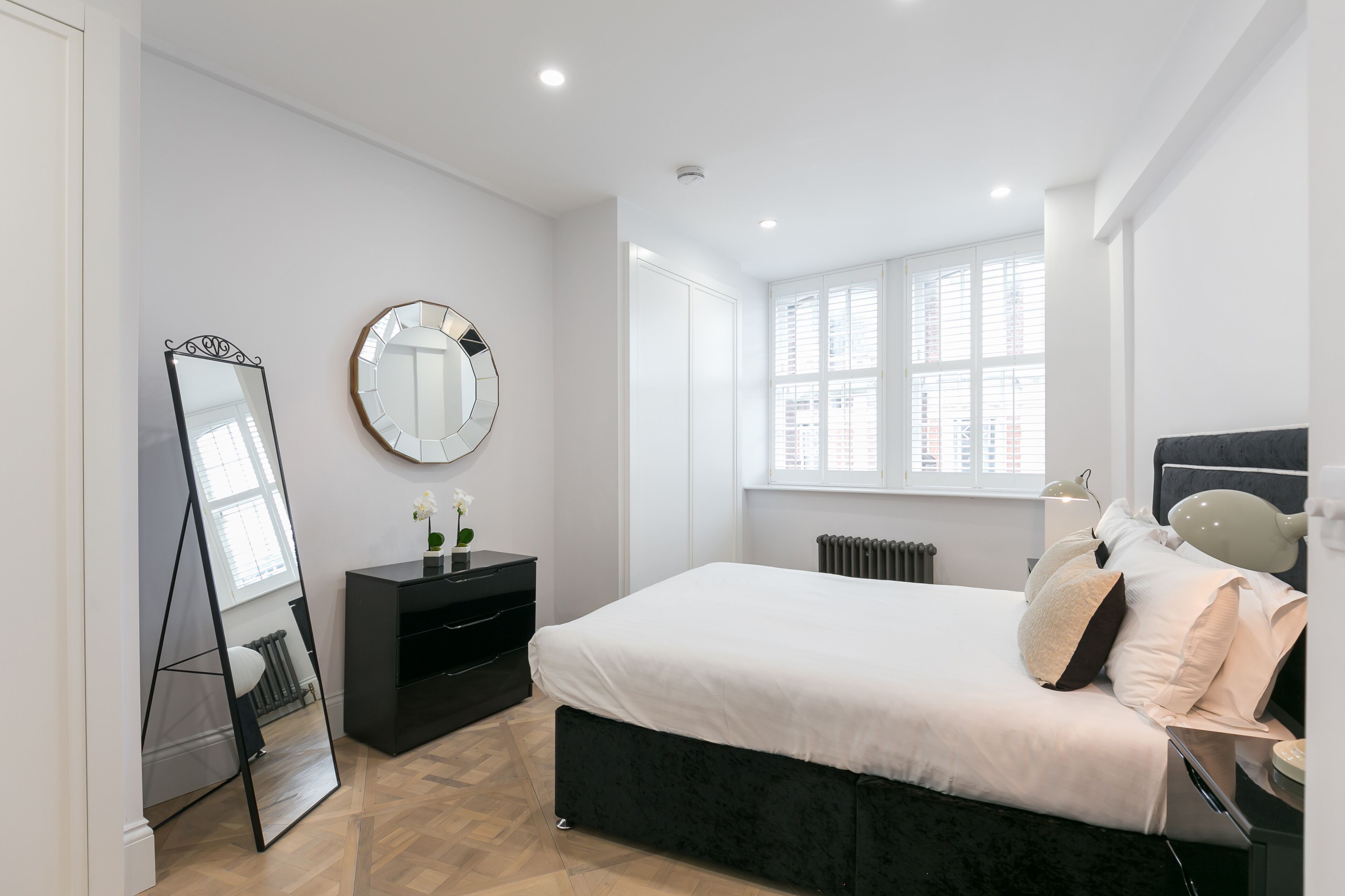 Une chambre moderne avec un grand lit, une commode, un miroir et une fenêtre avec des stores.