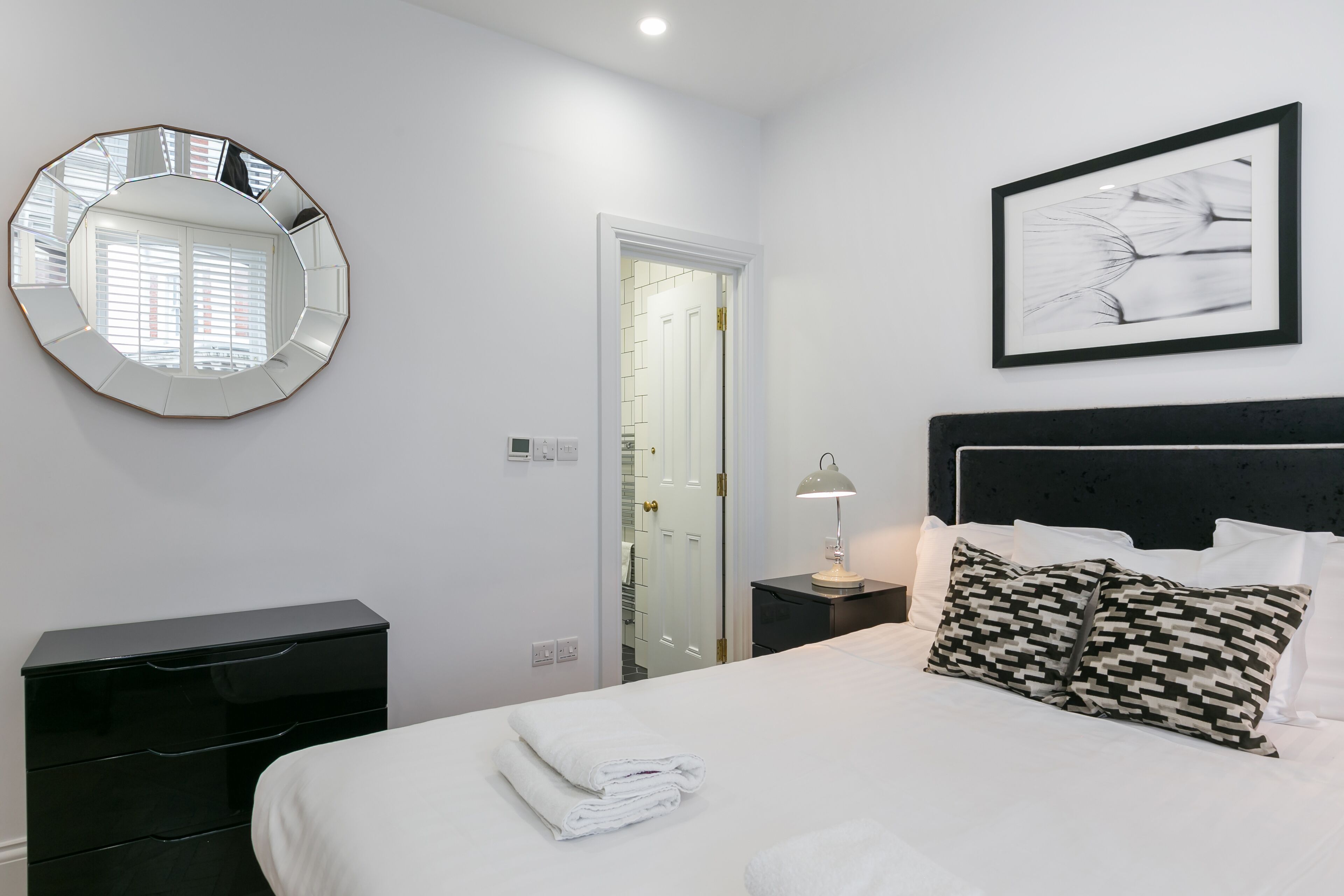 Una habitación de hotel con una cama, una mesita de noche con lámpara, un aparador negro, un espejo redondo y un cuadro en la pared.