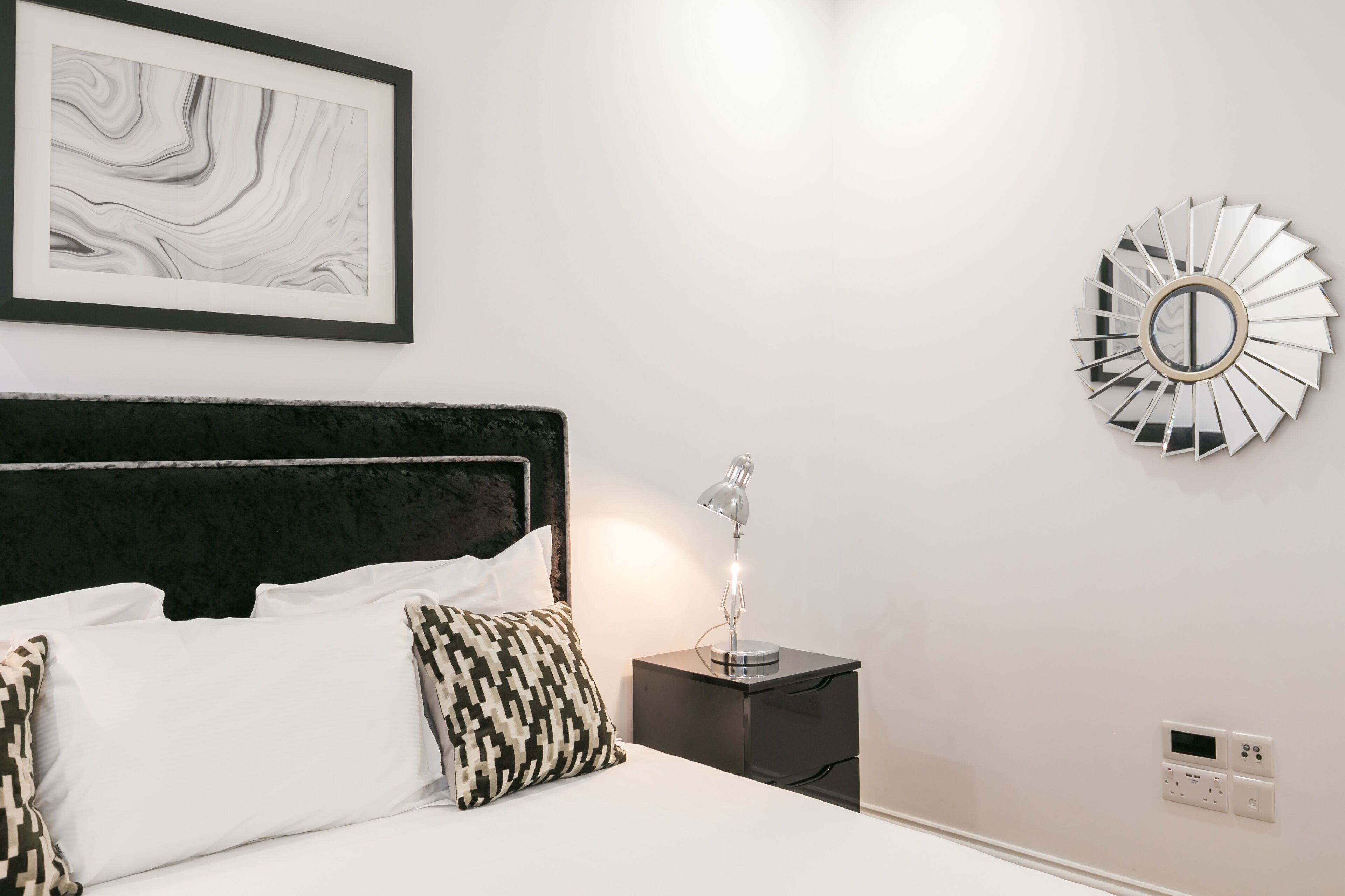 Una cama bien hecha con sábanas blancas y almohadas estampadas, un cabecero negro, una mesita de noche con una lámpara, una obra de arte abstracta enmarcada y un espejo decorativo en la pared.
