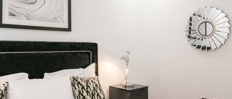 Una cama bien hecha con sábanas blancas y almohadas estampadas, un cabecero negro, una mesita de noche con una lámpara, una obra de arte abstracta enmarcada y un espejo decorativo en la pared.