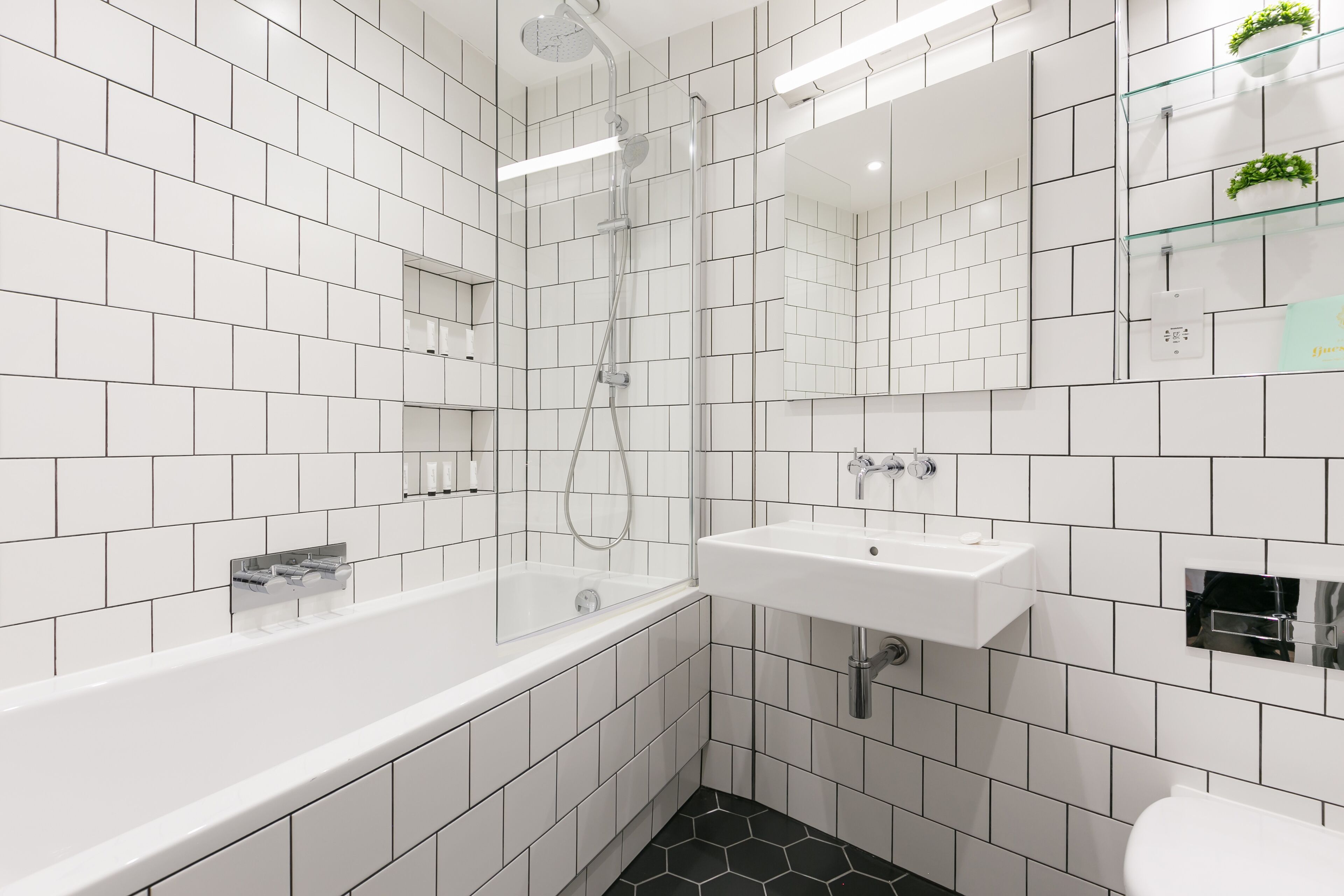 A modern bathroom with white tiles, a glass-enclosed bathtub, a white sink, and a mirror with a glass shelf above it.