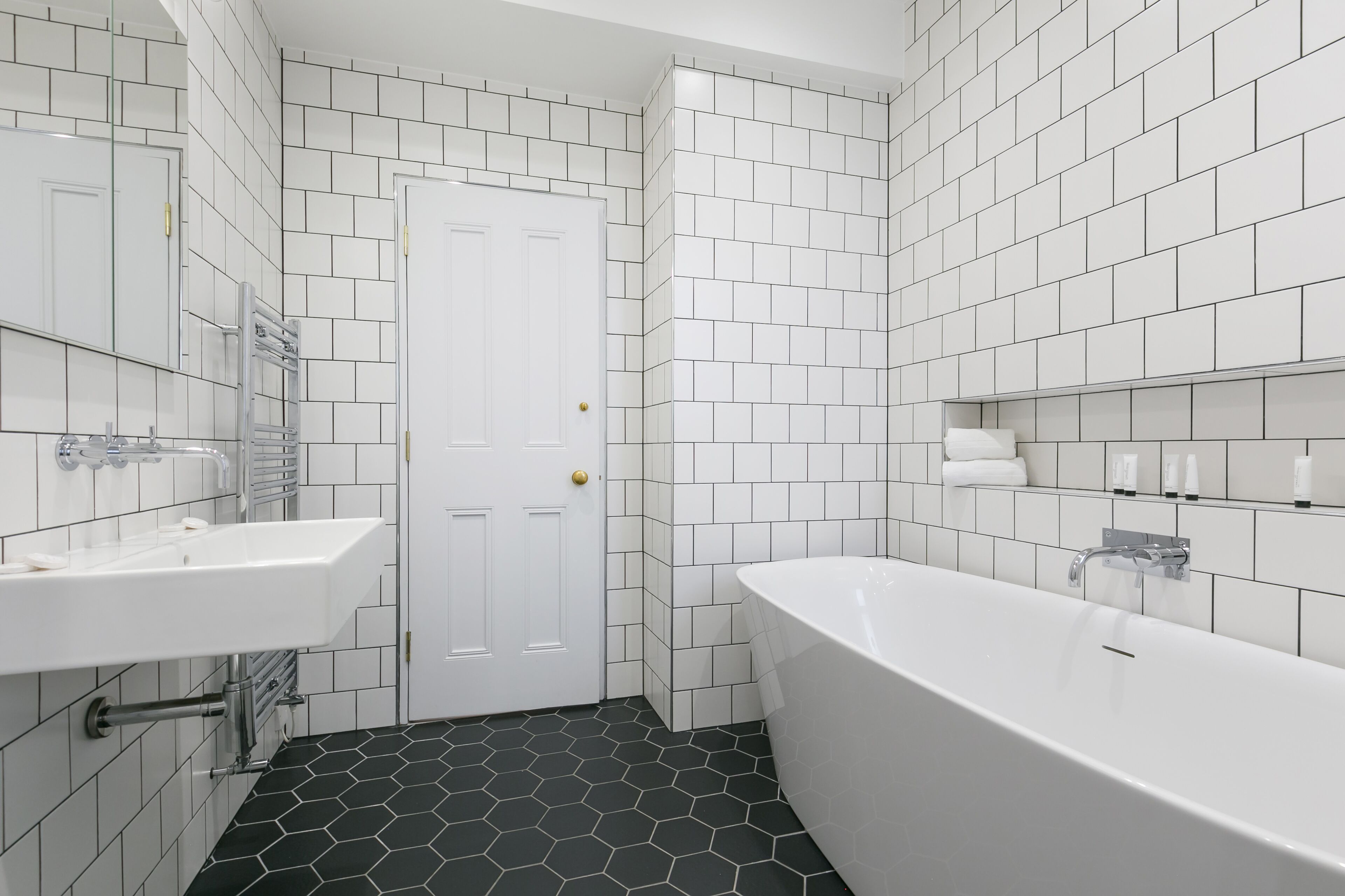 A modern bathroom with white tiled walls, a black hexagonal floor, a white bathtub, and a double sink.