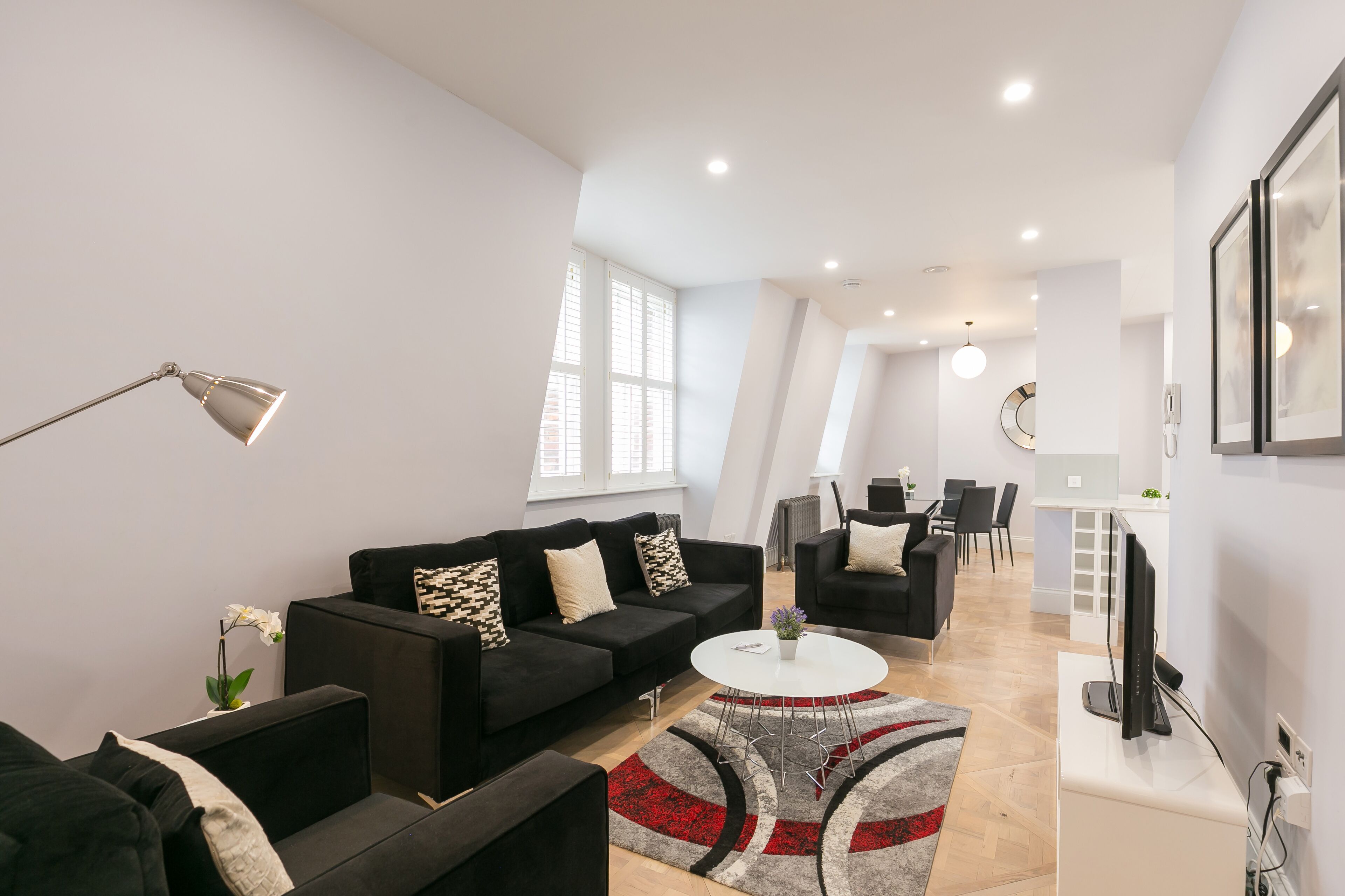 Un salon moderne avec un ensemble de canapés noirs, une table basse blanche et un tapis rouge et gris. On aperçoit à l’arrière-plan un coin repas avec une table et des chaises blanches.