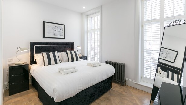 A modern bedroom with a large bed, a nightstand, a bedside table, a radiator, and a window with blinds.