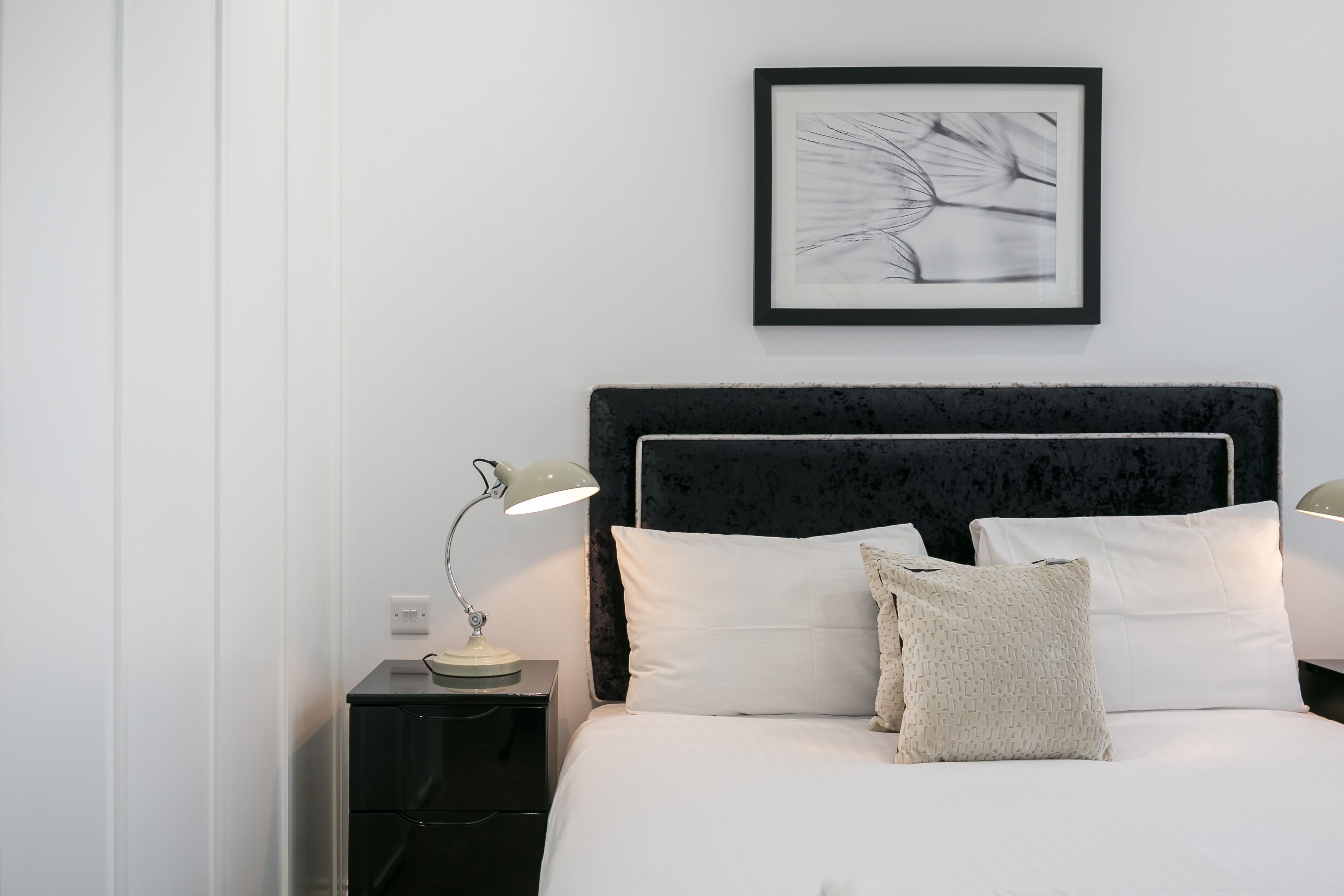 A neatly made bed with white linens and two pillows, a black headboard, a framed artwork on the wall, and a bedside lamp on a black nightstand.