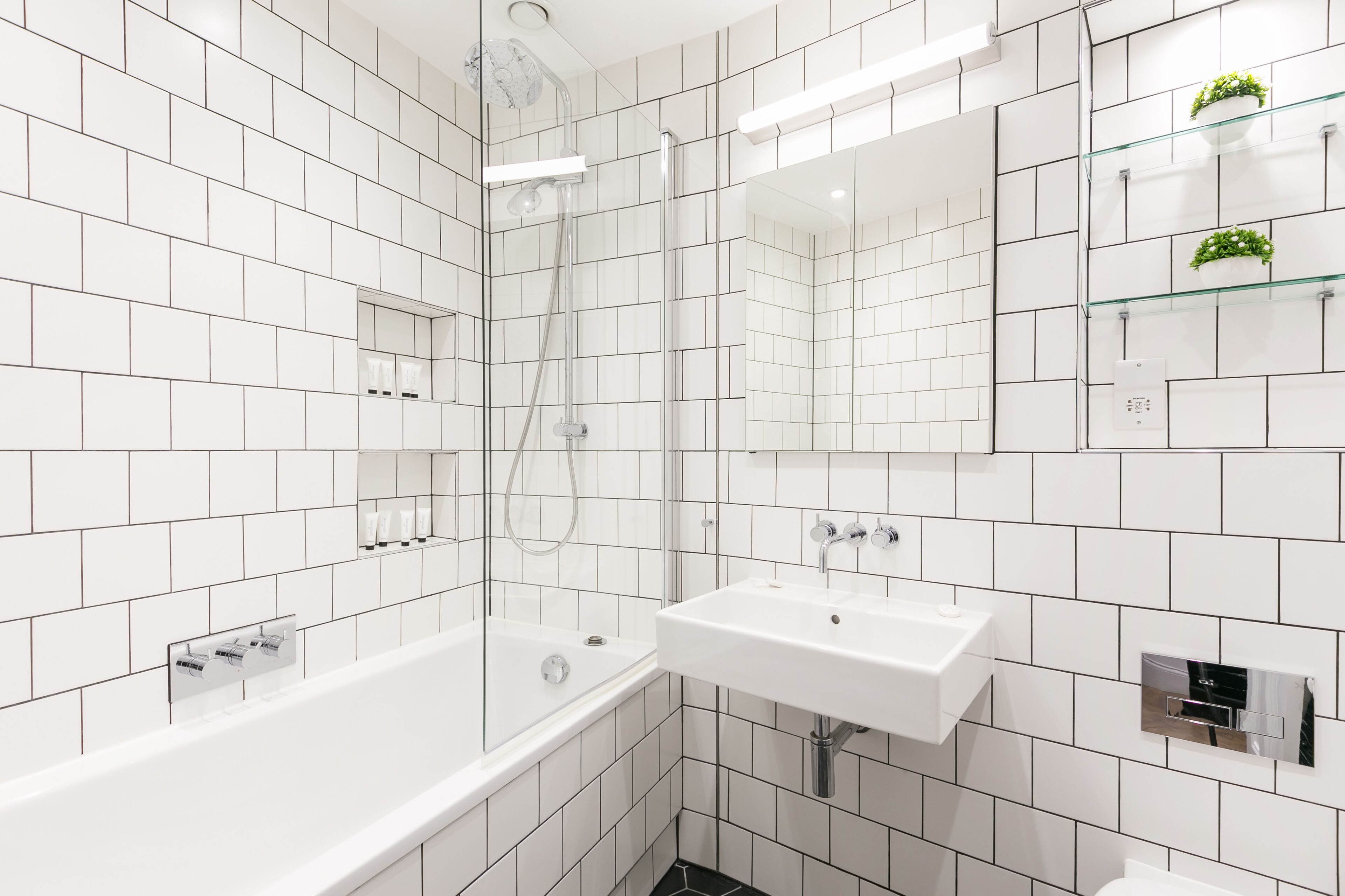 A modern bathroom with white tiles, a glass-enclosed shower, a white sink, and a mirror above it.