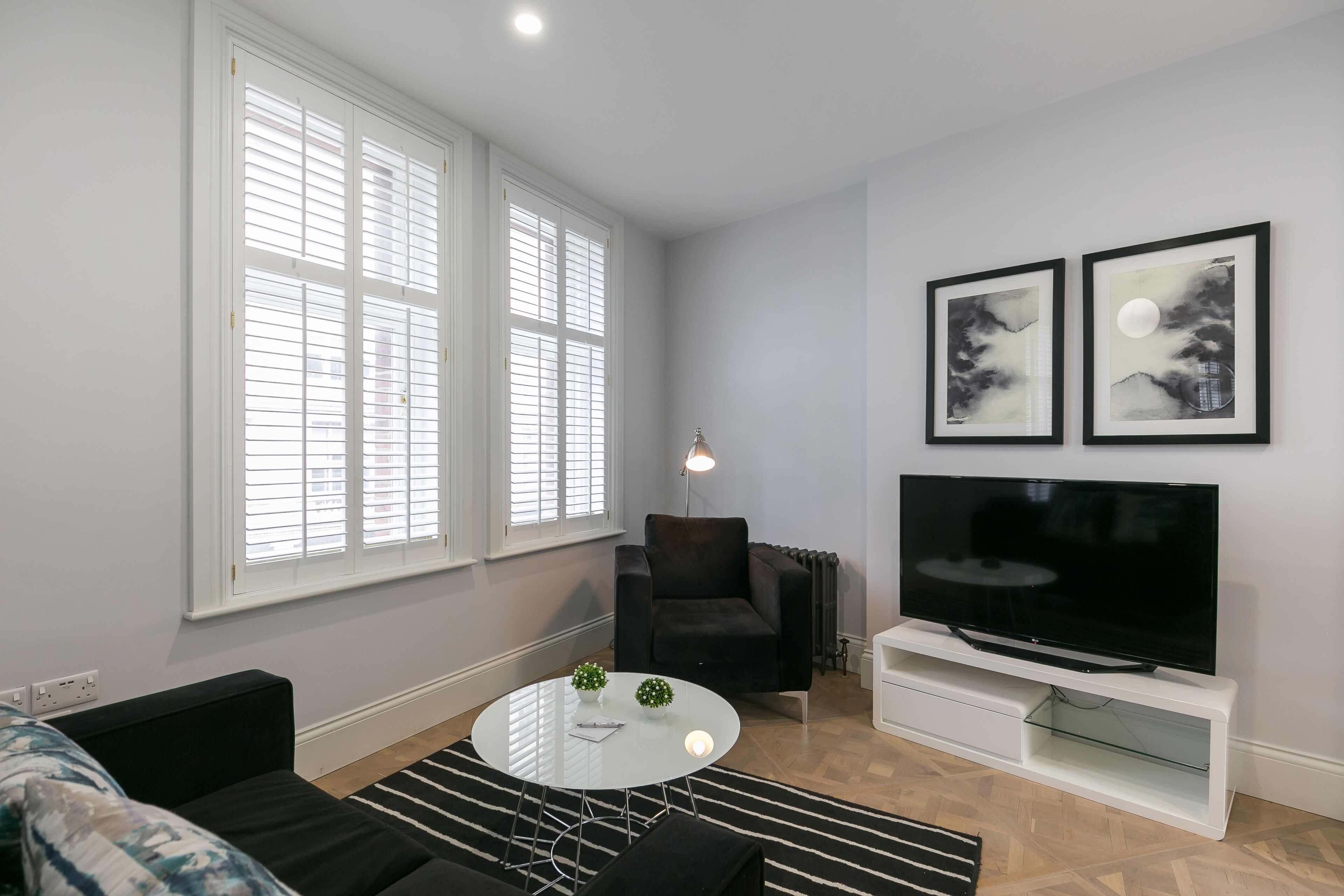 Una sala de estar moderna con un sofá negro, una mesa de centro blanca y un televisor sobre un mueble blanco. Hay dos cuadros en la pared y una lámpara de pie.