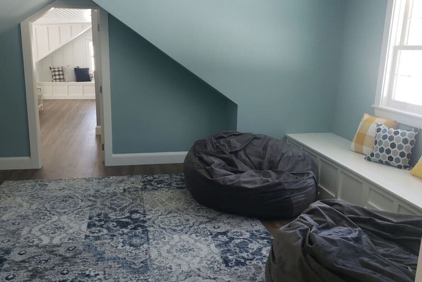 A room with a sloped ceiling, a patterned rug, a bean bag chair, and a window with decorative pillows.