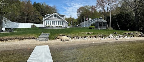 A clear view of a house with a large porch, a grassy area, and a body of water in the foreground.