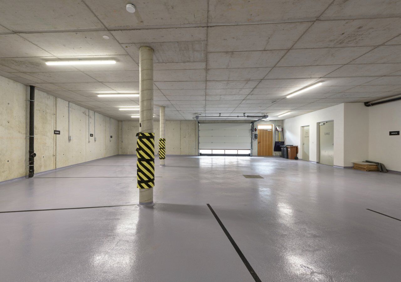 A spacious indoor parking area with concrete floors and walls, featuring a central pillar with yellow and black stripes, and a garage door at the far end.